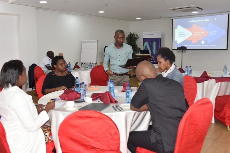 Participants sat around a table engaging in discussion during our 4 Billion Conversations Consultative event in Nairobi, Kenya. Paul from CLEAR Global is stood speaking to the group.