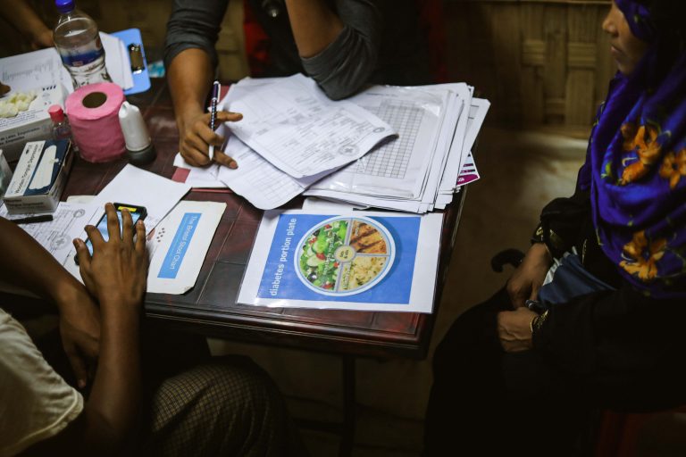 Rohingya woman and staff sit around a table covered in paperwork and a 'diabetes food plate' brochure