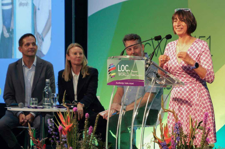 An image of Aimee Ansari, CEO Clear Global giving a speech at LocWorld 51 and 3 people sitting behind listening